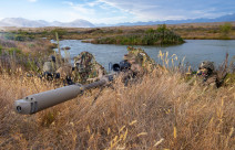 New Zealand Army soldiers lie in the grass around a lake area with the M107A1 Anti-Materiel Rifle. The backdrop shows the mountainous ranges around Tekapo Military Training Area. The grass is dry and its a sunny day with some cloud. 