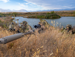 New Zealand Army soldiers lie in the grass around a lake area with the M107A1 Anti-Materiel Rifle. The backdrop shows the mountainous ranges around Tekapo Military Training Area. The grass is dry and its a sunny day with some cloud.