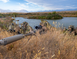 New Zealand Army soldiers lie in the grass around a lake area with the M107A1 Anti-Materiel Rifle. The backdrop shows the mountainous ranges around Tekapo Military Training Area. The grass is dry and its a sunny day with some cloud.