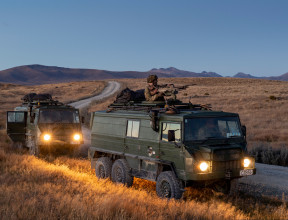 Two New Zealand Light Operational Vehicle (NZLOV) and soldiers in the evening light with their lights on. They are parked on the side of the gravel road and in the background you can see hills around the Waiouru Military Training Area