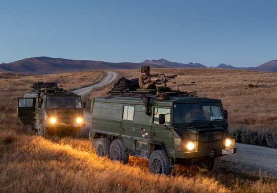 Two New Zealand Light Operational Vehicle (NZLOV) and soldiers in the evening light with their lights on. They are parked on the side of the gravel road and in the background you can see hills around the Waiouru Military Training Area
