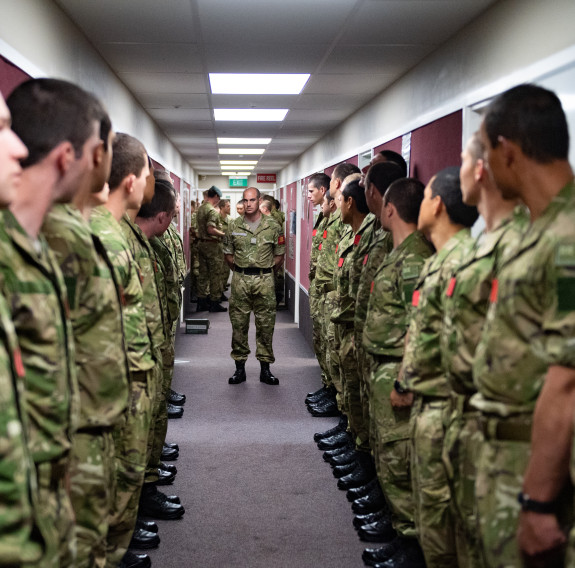 New Zealand Army soldiers stand in a corridor a row of people on each side. One solider stands in the middle of the corridor. 