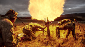 New Zealand Army soldiers fire a 81mm mortar round at dusk. One soldier stands back with looking at a piece of paper in lefthand side of the image. Two soldiers are near the mortar, heads down. Flames are shown coming out of the mortar which create the co