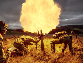 New Zealand Army soldiers fire a 81mm mortar round at dusk. One soldier stands back with looking at a piece of paper in lefthand side of the image. Two soldiers are near the mortar, heads down. Flames are shown coming out of the mortar which create the co