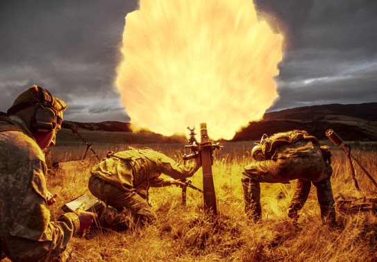 New Zealand Army soldiers fire a 81mm mortar round at dusk. One soldier stands back with looking at a piece of paper in lefthand side of the image. Two soldiers are near the mortar, heads down. Flames are shown coming out of the mortar which create the co