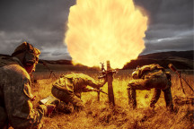 New Zealand Army soldiers fire a 81mm mortar round at dusk. One soldier stands back with looking at a piece of paper in lefthand side of the image. Two soldiers are near the mortar, heads down. Flames are shown coming out of the mortar which create the co