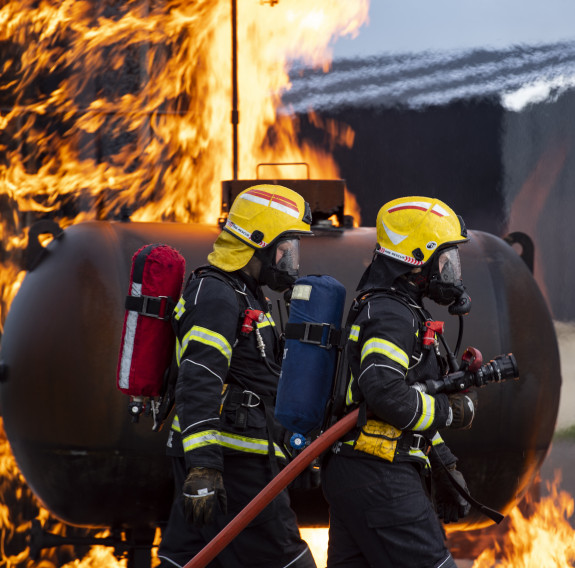 Two NZDF firefighters during a training activity. Both are in full breathing apparatus and one is carrying a hose. There are flames around them.