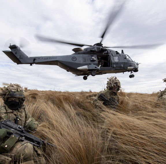 New Zealand Army soldiers sit in the tussock grass within the Waiouru Military Training Area after being dropped off by an NH90 helicopter (seen behind them flying away). 