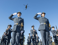 Two Royal New Zealand Air Force personnel salute during a graduation. Many other Air Force personnel stand around them and an NH90 helicopter flies overhead. It's a blue sky day and the image is taken from the ground looking up.
