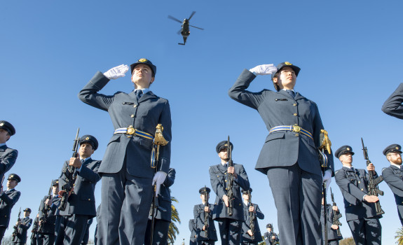 Two Royal New Zealand Air Force personnel salute during a graduation. Many other Air Force personnel stand around them and an NH90 helicopter flies overhead. It's a blue sky day and the image is taken from the ground looking up.