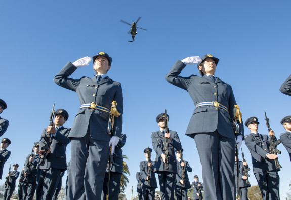 Two Royal New Zealand Air Force personnel salute during a graduation. Many other Air Force personnel stand around them and an NH90 helicopter flies overhead. It's a blue sky day and the image is taken from the ground looking up.
