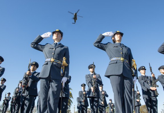 Two Royal New Zealand Air Force personnel salute during a graduation. Many other Air Force personnel stand around them and an NH90 helicopter flies overhead. It's a blue sky day and the image is taken from the ground looking up.