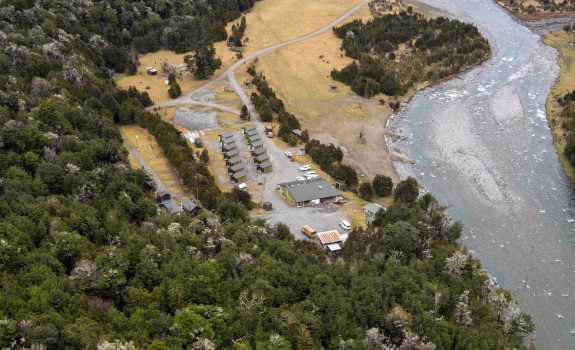 An aerial view of Dip Flat. This camp consists of one large building, 14 cabins and four other smaller buildings. The camp is located next to a river on the right and otherwise is surrounded by trees.