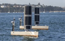 Two USVs painted grey sail in the water. The hull is grey and there is a large rectangle that stands up like a sail but is made of solar pannels.
