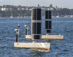 Two USVs painted grey sail in the water. The hull is grey and there is a large rectangle that stands up like a sail but is made of solar pannels.
