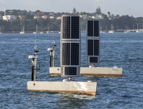 Two USVs painted grey sail in the water. The hull is grey and there is a large rectangle that stands up like a sail but is made of solar pannels.