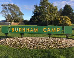 A green and yellow sign that says 'Burnham Camp' is fixed in the ground. In the background you see trees and blue sky
