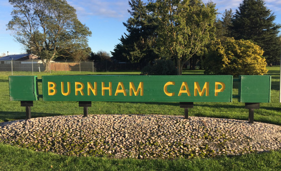 A green and yellow sign that says 'Burnham Camp' is fixed in the ground. In the background you see trees and blue sky