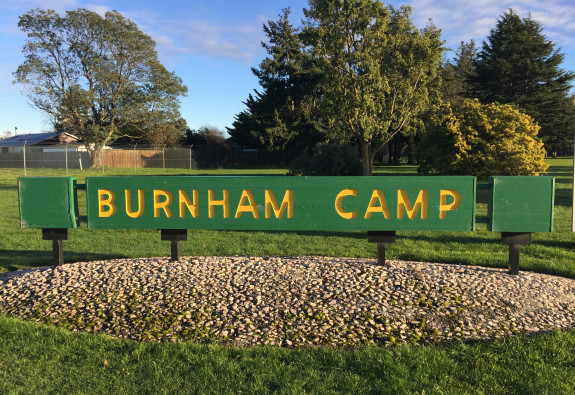A green and yellow sign that says 'Burnham Camp' is fixed in the ground. In the background you see trees and blue sky