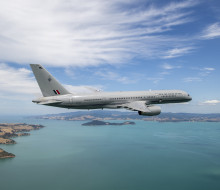 A Royal New Zealand Air Force Boeing 757-2K2 aircraft flies over the ocean but in the frame you can also see parts of land.