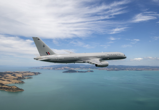 A Royal New Zealand Air Force Boeing 757-2K2 aircraft flies over the ocean but in the frame you can also see parts of land.