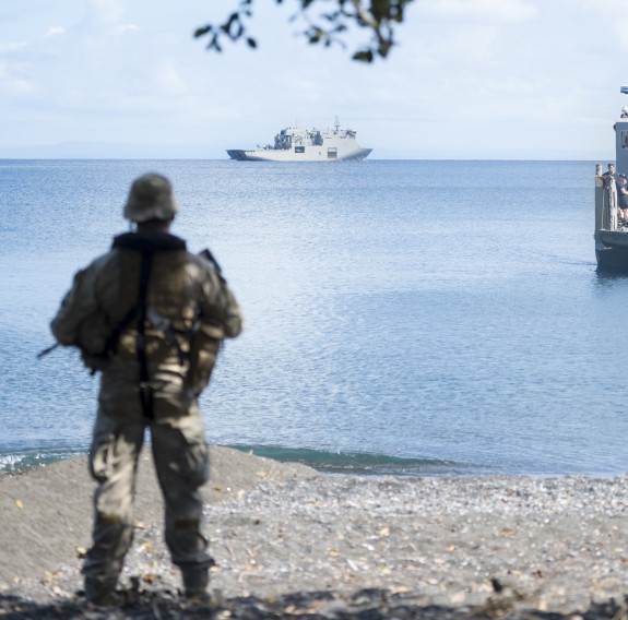 A New Zealand Army soldier watches from the shore as a Royal New Zealand Navy Landing Craft Mechanised (LCM) is sent from HMNZS Canterbury. The image is framed with branches from a tree. 