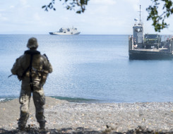 A New Zealand Army soldier watches from the shore as a Royal New Zealand Navy Landing Craft Mechanised (LCM) is sent from HMNZS Canterbury. The image is framed with branches from a tree. 