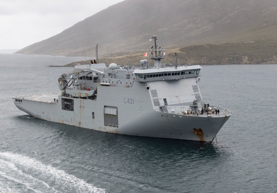 HMNZS Canterbury sailing in the ocean, a Rigid Hull Inflatable Boat (RHIB) moves toward the ship from the bottom of the image. The day looks dreary and cloudy.