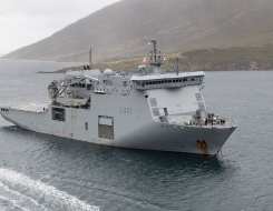 HMNZS Canterbury sailing in the ocean, a Rigid Hull Inflatable Boat (RHIB) moves toward the ship from the bottom of the image. The day looks dreary and cloudy.