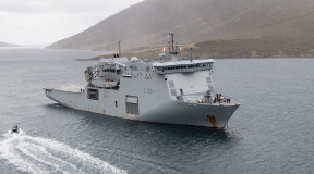 HMNZS Canterbury sailing in the ocean, a Rigid Hull Inflatable Boat (RHIB) moves toward the ship from the bottom of the image. The day looks dreary and cloudy.