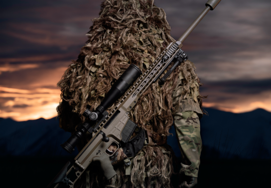 A soldier stands with their back facing the camera and the MRAD Sniper Rifle on their back. The background of the image has the sun setting and is very dark with some yellow/orange sky