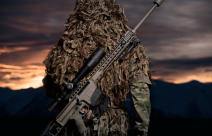 A soldier stands with their back facing the camera and the MRAD Sniper Rifle on their back. The background of the image has the sun setting and is very dark with some yellow/orange sky