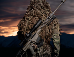 A soldier stands with their back facing the camera and the MRAD Sniper Rifle on their back. The background of the image has the sun setting and is very dark with some yellow/orange sky