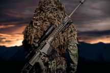 A soldier stands with their back facing the camera and the MRAD Sniper Rifle on their back. The background of the image has the sun setting and is very dark with some yellow/orange sky