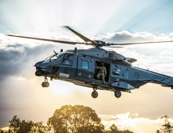 An NH90 helicopter flies in the sky with the helicopter loadmaster looking out the door to check the ground before landing. The sun is streaming behind the helicopter and cloud behind it. 