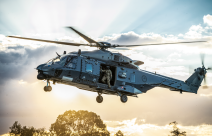 An NH90 helicopter flies in the sky with the helicopter loadmaster looking out the door to check the ground before landing. The sun is streaming behind the helicopter and cloud behind it. 
