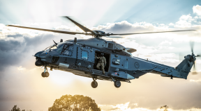 An NH90 helicopter flies in the sky with the helicopter loadmaster looking out the door to check the ground before landing. The sun is streaming behind the helicopter and cloud behind it. 