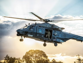 An NH90 helicopter flies in the sky with the helicopter loadmaster looking out the door to check the ground before landing. The sun is streaming behind the helicopter and cloud behind it. 