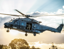 An NH90 helicopter flies in the sky with the helicopter loadmaster looking out the door to check the ground before landing. The sun is streaming behind the helicopter and cloud behind it. 