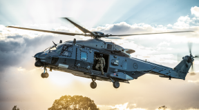 An NH90 helicopter flies in the sky with the helicopter loadmaster looking out the door to check the ground before landing. The sun is streaming behind the helicopter and cloud behind it. 