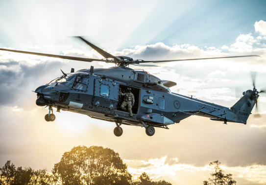 An NH90 helicopter flies in the sky with the helicopter loadmaster looking out the door to check the ground before landing. The sun is streaming behind the helicopter and cloud behind it. 