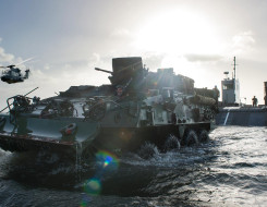 A New Zealand Army Light Armoured Vehicle (NZLAV) moves through the water after coming off a Landing Craft Mechanism (LCM). To the left is an NH90 helicopter and in the far distance, behind the LCM is HMNZS Canterbury. It's a sunny day and the sun is cre