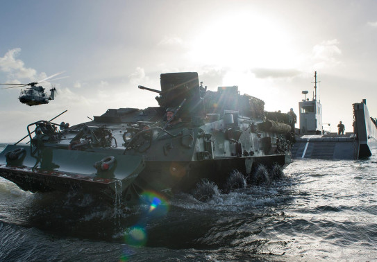 A New Zealand Army Light Armoured Vehicle (NZLAV) moves through the water after coming off a Landing Craft Mechanism (LCM). To the left is an NH90 helicopter and in the far distance, behind the LCM is HMNZS Canterbury.  It's a sunny day and the sun is cre