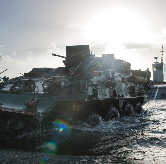 A New Zealand Army Light Armoured Vehicle (NZLAV) moves through the water after coming off a Landing Craft Mechanism (LCM). To the left is an NH90 helicopter and in the far distance, behind the LCM is HMNZS Canterbury.  It's a sunny day and the sun is cre