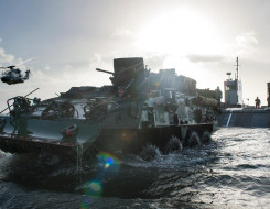 A New Zealand Army Light Armoured Vehicle (NZLAV) moves through the water after coming off a Landing Craft Mechanism (LCM). To the left is an NH90 helicopter and in the far distance, behind the LCM is HMNZS Canterbury.  It's a sunny day and the sun is cre