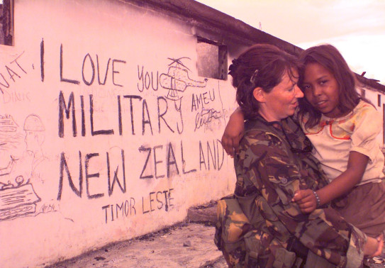 A New Zealand Army soldiers holds a young child in the right of the image. On the left is the wall of a building that reads "I love you military New Zealand Timor Leste - Ameu' with military drawings around it. The photo has a pink colouring to it. 
