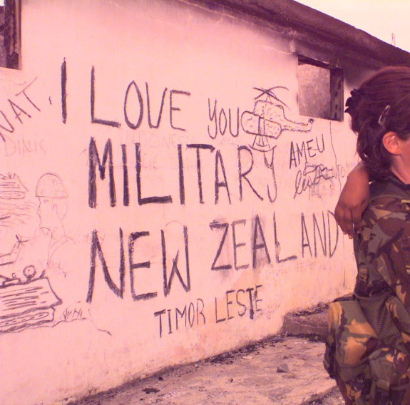 A New Zealand Army soldiers holds a young child in the right of the image. On the left is the wall of a building that reads "I love you military New Zealand Timor Leste - Ameu' with military drawings around it. The photo has a pink colouring to it. 