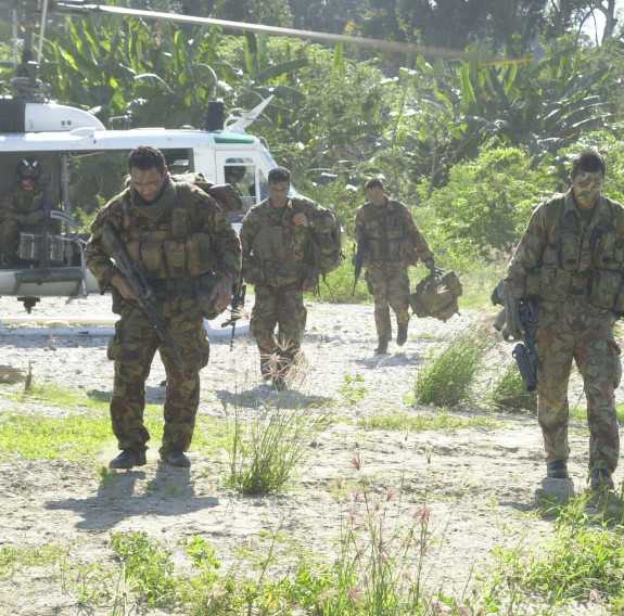 Four uniformed soldiers walk toward the camera after getting off a helicopter in a clearing within a jungle.
