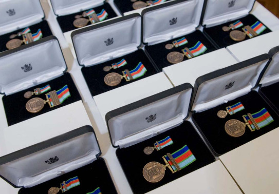 Many boxes of New Zealand Defence Service Medals. In each box is the medal, a bronze medal with an antique finish suspended from a dark blue, red, light blue and light green ribbon. 