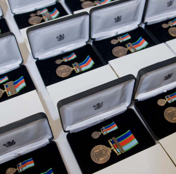 Many boxes of New Zealand Defence Service Medals. In each box is the medal, a bronze medal with an antique finish suspended from a dark blue, red, light blue and light green ribbon. 